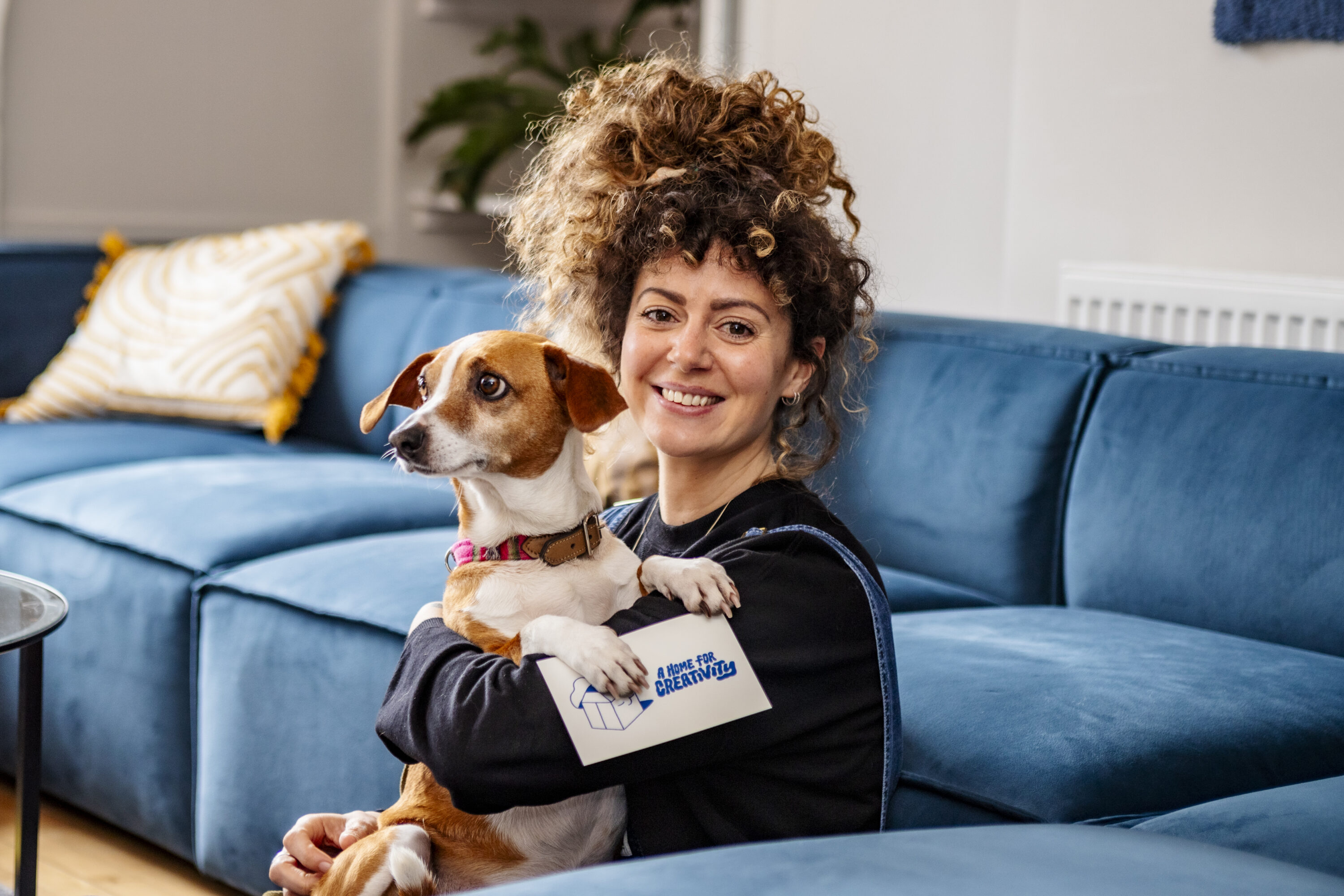 A smiling woman with curly brown hair sits on a blue couch, holding a small brown and white dog in her arms. She is wearing a black sweater and denim overalls. The dog has a pink collar and looks off to the side. The woman holds a white card with the text "A HOME FOR CREATIVITY" and a logo of a house in blue. The background features a blue couch with yellow and white patterned pillows and a potted plant on a shelf.