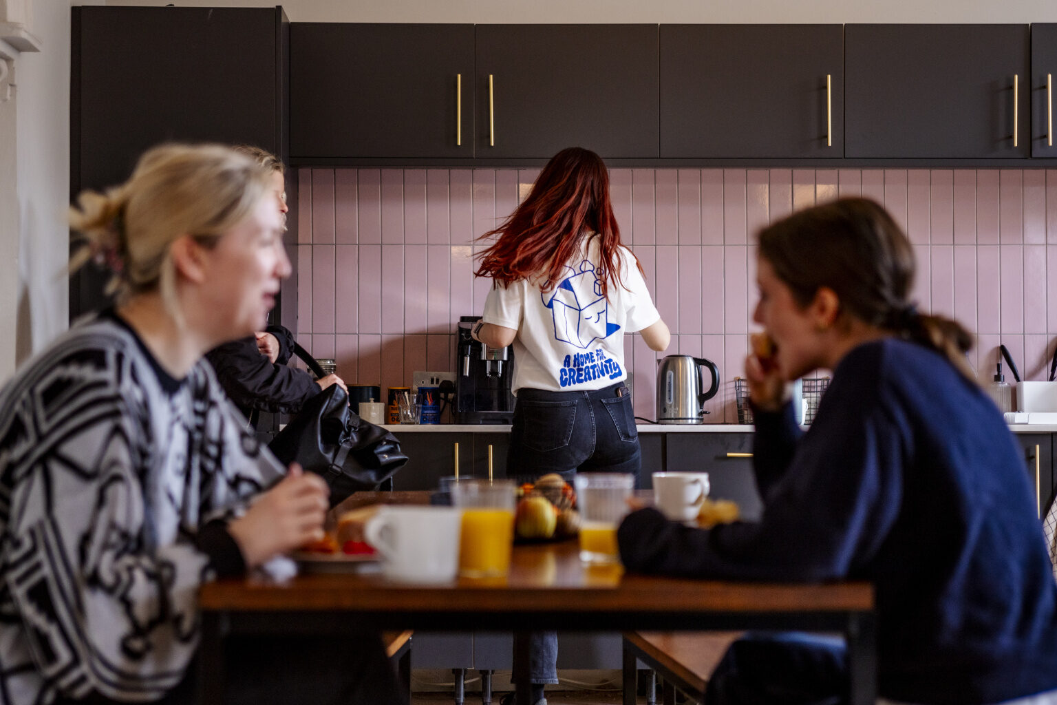 Gather Round photography. Photo shows 2 women sat at a table chatting in the foreground, with another woman in the kitchen behind. Interior looks modern, with pale pink tiles on the wall.
