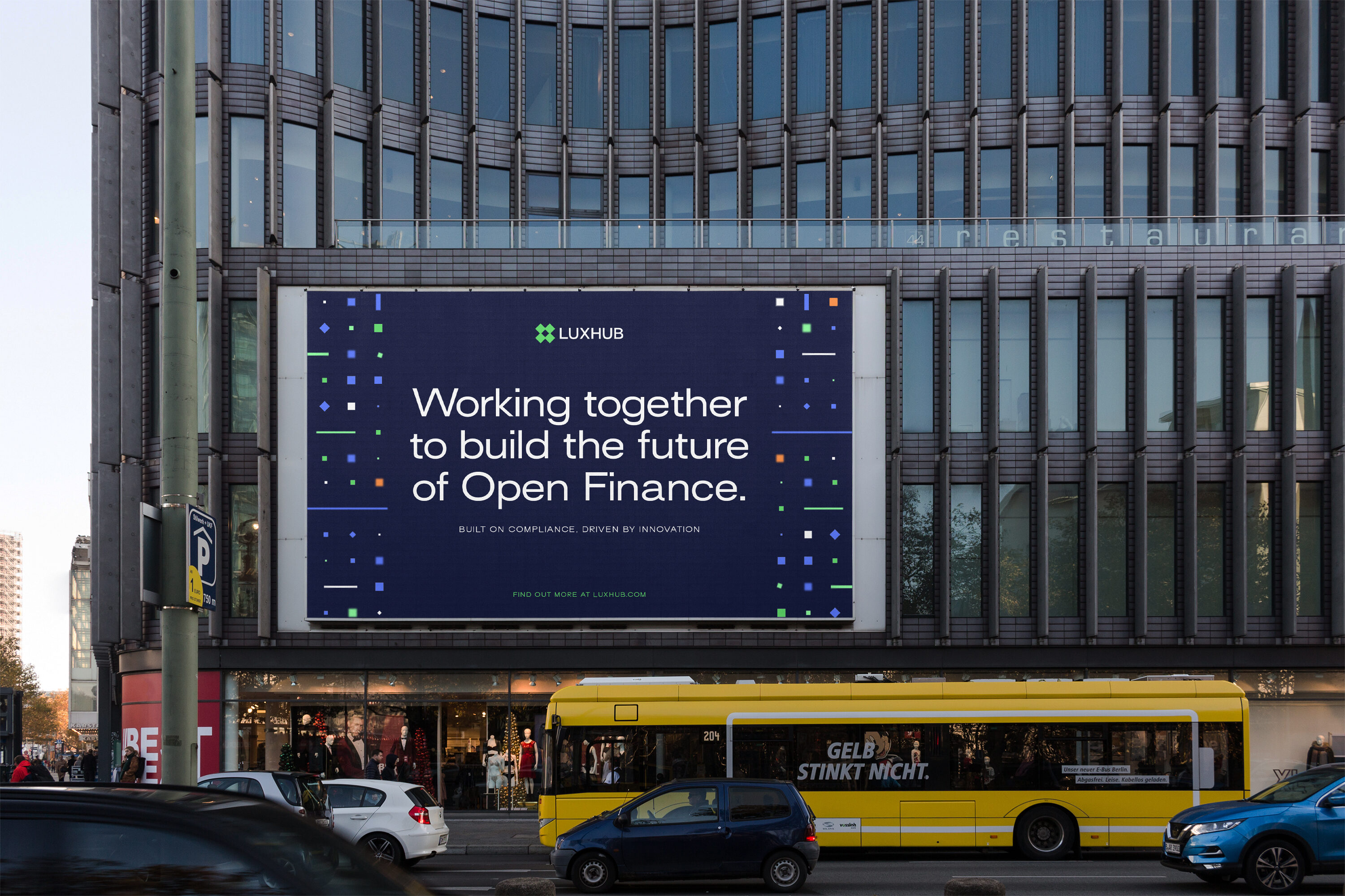 Luxhub billboard mock up. A dark blue 16:9 billboard is positioned on the side of a building within an urban scene. The building has a glass and curved brick facade, with vertical slats. There is a shopfront below and a road with traffic passing by, including a yellow bus. The billboard features the prominent headline 'Working together to build the future of Open Finance' in the centre in white text across three lines. The Luxhub logo is shown above this. The Luxhub logomark is represented by a green 'X', created by four squares, all connected in the centre by an interior negative space. A white all caps wordmark is to its right. Secondary copy is written underneath in smaller white all caps text 'Built on compliance. Driven by innovation' with a green all caps text call to action 'Find out more at Luxhub.com'. The left and right hand sections of the billboard feature a grid-like pattern made of different sized white, blue, green, and occasional orange squares, rectangles and dots.