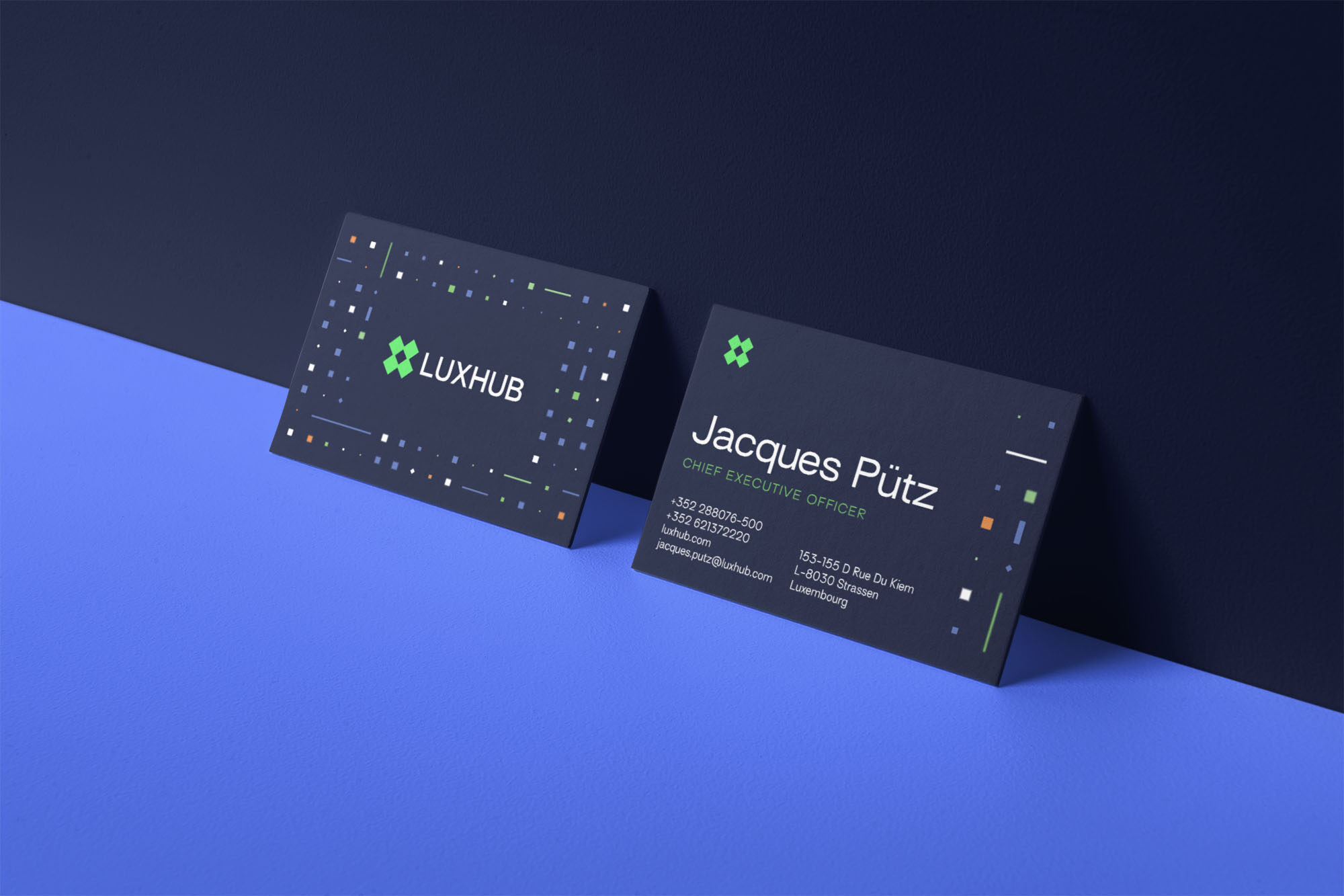 Luxhub business card mock up. Two dark blue business cards on a blue surface lean against an angled dark blue wall. The business cards are predominantly dark blue in colour. The left card features the Luxhub logomark in green, with a white all caps wordmark to its right. The Luxhub logomark is represented by a green 'X', created by four squares, all connected in the centre by an interior negative space. Around the outer half of the card there is grid-like pattern made of different sized white, blue, green, and occasional orange squares, rectangles and dots. The card on the right has the Luxhub green logomark in the top left corner. Along the centre of the card the name 'Jacques Pütz' is written in white. Underneath this in smaller green all caps text is the title 'Chief Executive Officer', and then two columns of smaller white text details contact information in two columns. The grid-like pattern described above runs along the right side of the card.