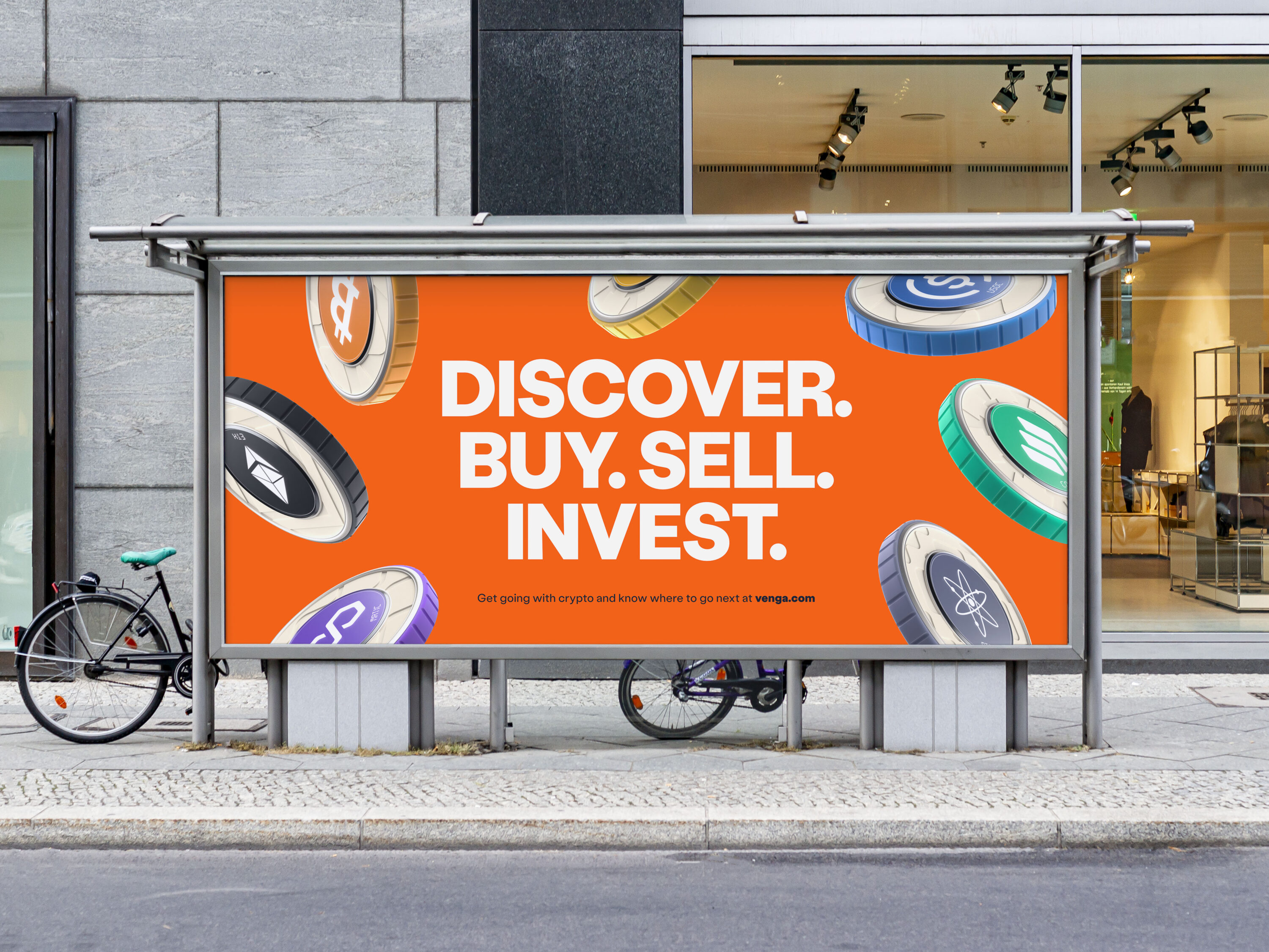 Venga billboard mock up. The billboard shows various crypto coins. Each coin type has a central circular colour - including orange, aqua, blue, purple and black - which is also repeated along its edge. The centre circle contains the coin's icon and shortened name in white. Each coin type has its own subtle textured brand pattern. The coins float in front of an orange background. The headline reads 'Discover. Buy. Sell. Invest' in white bold all caps. There is a small call to action line with a weblink to venga.com. The billboard is placed within a street scene, there is a concrete wall and shopfront behind it. Two bikes in partial view lean against it.