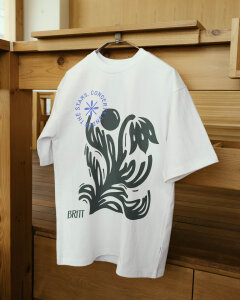Image shows a mockup of the Britt fauna logo on a tshirt. The shirt is white, with a dark green motif printed on, This is hanging on a hanger with a wooden door.