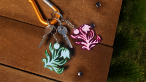 Image shows branded Britt merch as keyrings, using the fauna logo. There are two keyrings pictured, on in a green and one in a complimentary pink. These are laid on a wooden table, with grass in background.