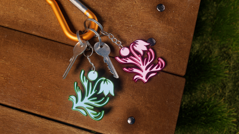 Image shows branded Britt merch as keyrings, using the fauna logo. There are two keyrings pictured, on in a green and one in a complimentary pink. These are laid on a wooden table, with grass in background.